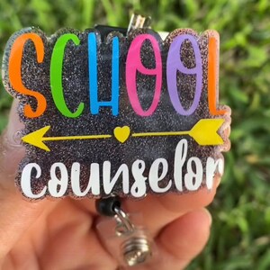 Może przedstawiać: Czarny błyszczący uchwyt na identyfikator z napisem "SCHOOL COUNSELOR" w kolorach tęczy i ż&oacute;łtą strzałką. Uchwyt na identyfikator ma czarną, chowaną szpulę.
