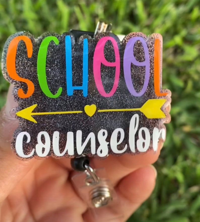 Może przedstawiać: Czarny błyszczący uchwyt na identyfikator z napisem "SCHOOL COUNSELOR" w kolorach tęczy i ż&oacute;łtą strzałką. Uchwyt na identyfikator ma czarną, chowaną szpulę.