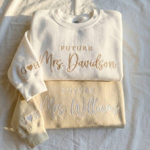 May include: Two cream-colored sweatshirts with embroidered text. The top sweatshirt reads "FUTURE Mrs. Davidson" in gold thread, with initials and a heart on the sleeve. The bottom sweatshirt is a light yellow color and reads "FUTURE Mrs. Williams" in white thread.