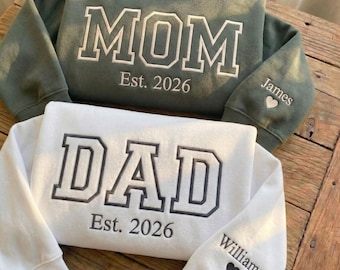 May include: An olive green sweatshirt with white embroidered text reading MOM Est. 2026 and a white sweatshirt with dark gray embroidered text reading DAD Est. 2026. The sleeves feature embroidered names James and Williams with small heart icons.