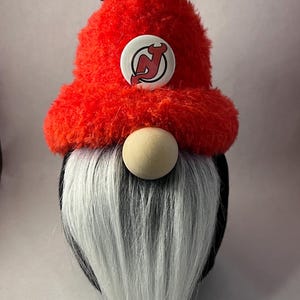 May include: A red and black gnome with a white beard. The gnome is wearing a red hat with a black and white pom-pom and a New Jersey Devils logo.