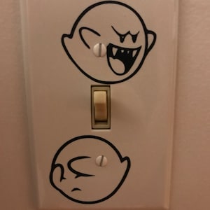 May include: A white light switch with two black and white ghost stickers on it. The ghosts are from the video game Super Mario Bros.