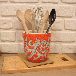 Puede incluir: Porta utensilios de cerámica naranja con un diseño floral blanco. El porta utensilios está lleno de cucharas de madera y un batidor.