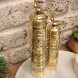May include: Two gold metal pepper grinders with intricate designs. The larger grinder has a handle on top. The grinders are sitting on a white marble surface.