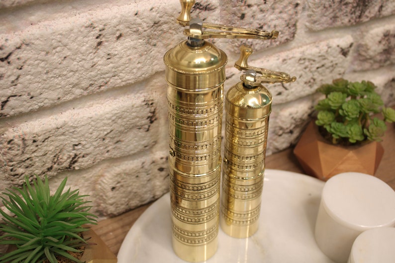 May include: Two gold metal pepper grinders with intricate designs. The larger grinder has a handle on top. The grinders are sitting on a white marble surface.