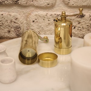 May include: A brass pepper grinder with a crank handle, a brass lid, and a brass base. The grinder is open and the inside is visible. The grinder is on a white marble surface with four white ceramic containers.