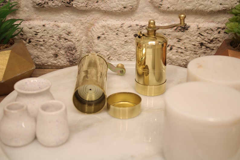May include: A brass pepper grinder with a crank handle, a brass lid, and a brass base. The grinder is open and the inside is visible. The grinder is on a white marble surface with four white ceramic containers.