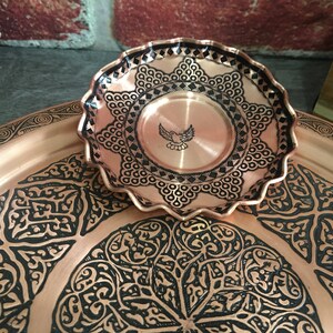 May include: A small, round, scalloped-edge copper dish featuring a central engraved bird motif and intricate black geometric patterns, resting atop a larger, matching copper tray decorated with elaborate, repeating black floral-inspired designs.