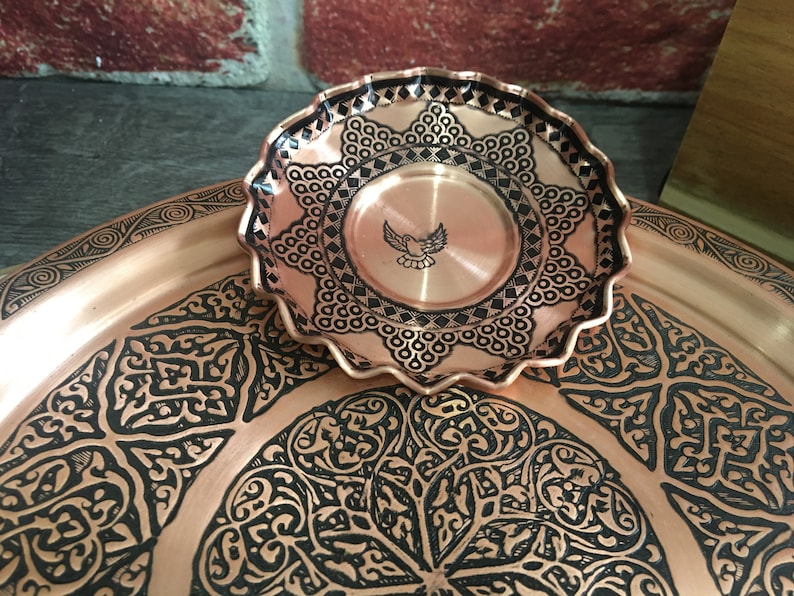 May include: A small, round, scalloped-edge copper dish featuring a central engraved bird motif and intricate black geometric patterns, resting atop a larger, matching copper tray decorated with elaborate, repeating black floral-inspired designs.
