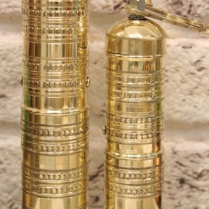 May include: Two gold metal pepper mills with intricate designs. The mills are made of brass and have a traditional style.