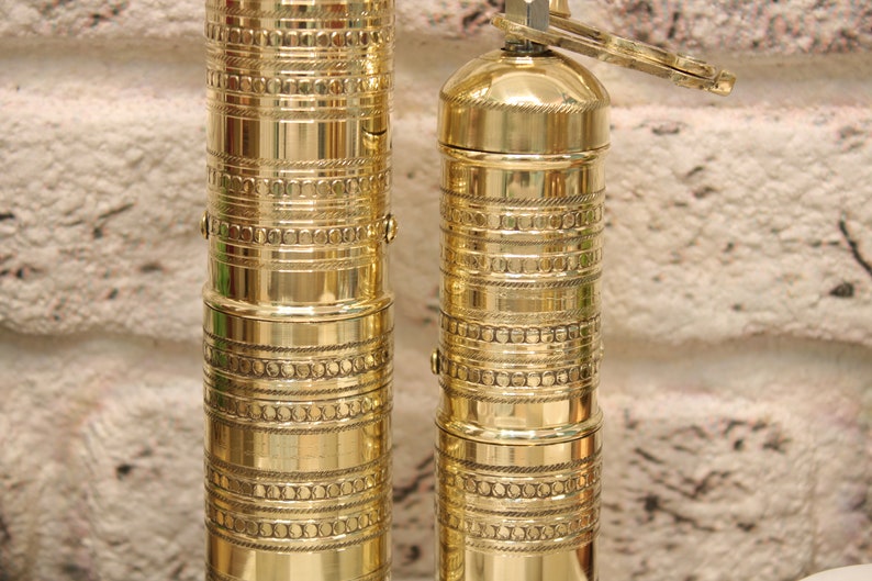 May include: Two gold metal pepper mills with intricate designs. The mills are made of brass and have a traditional style.