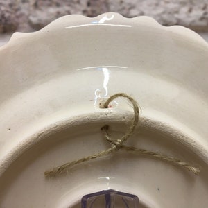 Puede incluir: La parte posterior de un plato de cer&aacute;mica blanco con un borde festoneado. Un trozo de cuerda est&aacute; atado a trav&eacute;s de un agujero en el plato para colgarlo.