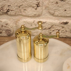 May include: Two gold metal pepper grinders with a crank handle on a white marble surface.