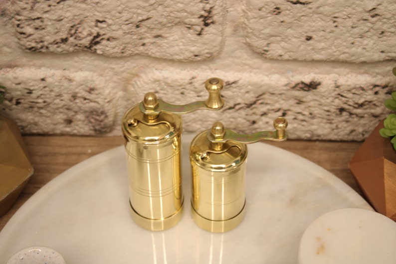 May include: Two gold metal pepper grinders with a crank handle on a white marble surface.