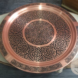 May include: A round copper tray featuring a polished metallic surface with intricate black etched patterns. The design includes a central circular motif surrounded by geometric shapes and a decorative border. A logo reads QUBICCA HANDICRAFT.