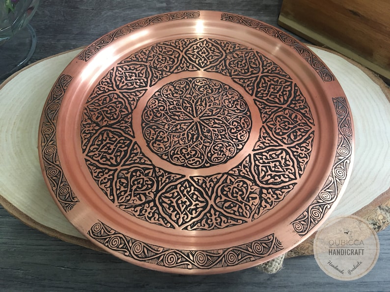 May include: A round copper tray featuring a polished metallic surface with intricate black etched patterns. The design includes a central circular motif surrounded by geometric shapes and a decorative border. A logo reads QUBICCA HANDICRAFT.