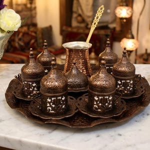 May include: A copper-toned coffee set featuring a central hammered pot with a long handle, surrounded by six lidded cups with intricate filigree metal casings over white ceramic inserts, all arranged on a matching round, embossed serving tray.