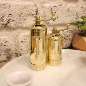 May include: Two gold metal pepper mills with a crank on top. The mills are sitting on a white marble surface.
