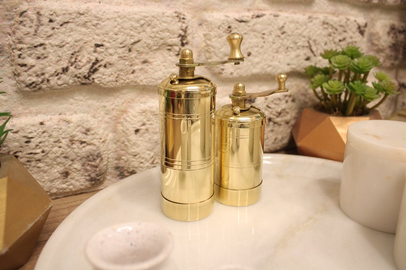 May include: Two gold metal pepper mills with a crank on top. The mills are sitting on a white marble surface.