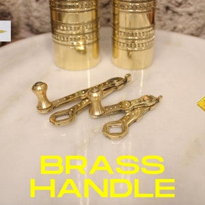 May include: Two gold-toned brass handles with a decorative design. The handles are on a white marble surface. The text "BRASS HANDLE" is written in yellow letters below the handles.