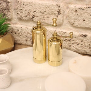 May include: Two gold metal pepper grinders with a crank handle on a white marble surface. The grinders are on a white marble tray with three small white ceramic pots.