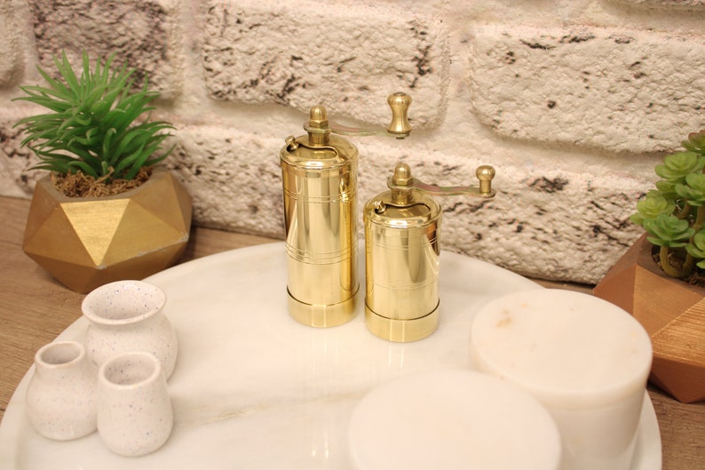 May include: Two gold metal pepper grinders with a crank handle on a white marble surface. The grinders are on a white marble tray with three small white ceramic pots.
