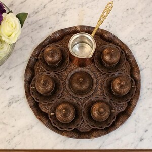 A set of six ornate copper coffee cups and saucers with intricate designs, a matching copper coffee pot, and a wooden tray. The cups and saucers are arranged in a circle around the coffee pot, and the tray is decorated with a floral pattern.