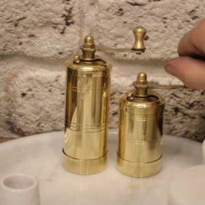 May include: Two gold metal pepper grinders with a hand turning the top of one of the grinders. The grinders are on a white marble surface.