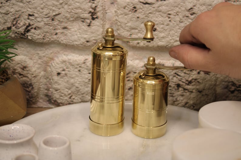 May include: Two gold metal pepper grinders with a hand turning the top of one of the grinders. The grinders are on a white marble surface.
