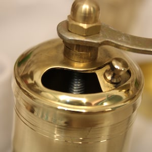 May include: A gold metal pepper grinder with a crank handle. The grinder has a small opening on the top for adding peppercorns.