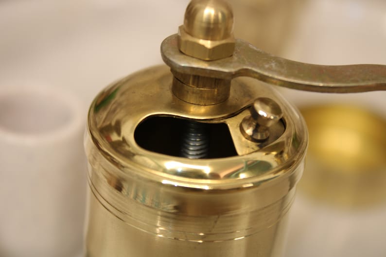 May include: A gold metal pepper grinder with a crank handle. The grinder has a small opening on the top for adding peppercorns.