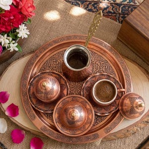 May include: A copper coffee set featuring a round tray with intricate etched patterns, a tall coffee pot with a long handle, two lidded containers, and a small cup filled with dark coffee, all displaying a consistent metallic finish.
