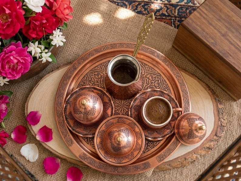 May include: A copper coffee set featuring a round tray with intricate etched patterns, a tall coffee pot with a long handle, two lidded containers, and a small cup filled with dark coffee, all displaying a consistent metallic finish.