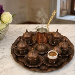 May include: A copper-coloured coffee serving set featuring a round tray, a central coffee pot with a long handle, and six small cups with matching lids and saucers. The metal surfaces are decorated with intricate embossed patterns throughout.