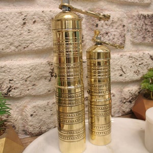 May include: Two gold metal pepper grinders with intricate designs. The larger grinder is on the left and the smaller grinder is on the right.