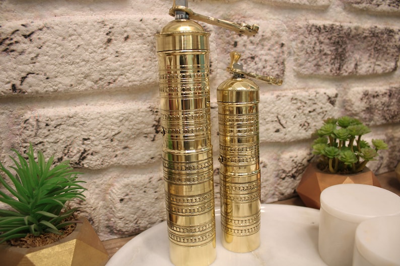 May include: Two gold metal pepper grinders with intricate designs. The larger grinder is on the left and the smaller grinder is on the right.