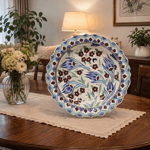 May include: A round ceramic decorative plate with a scalloped edge, featuring a hand-painted floral pattern with blue tulip motifs and small dark red flowers on a white background, framed by a blue and white petal-like border design.
