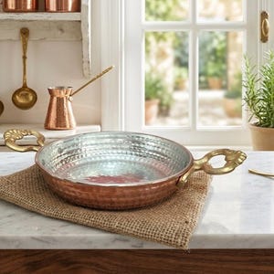 Puede incluir: Una sartén redonda y poco profunda de cobre martillado con un revestimiento interior plateado y dos asas de latón curvas y ornamentadas a los lados. El exterior presenta un acabado texturizado y con hoyuelos, con asas de diseño decorativo.