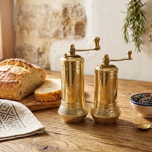 Puede incluir: Dos molinillos de especias cilíndricos de latón con manivelas manuales y detalles en relieve ornamentales alrededor de la base. Los molinillos presentan un acabado metálico pulido y están colocados en posición vertical con bandas horizontales.