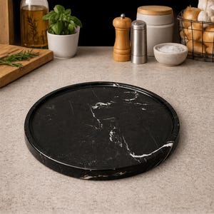 May include: A circular black marble tray with a raised edge and white veining patterns. The tray has a smooth, polished surface and is positioned on a light-colored countertop, showcasing its natural stone texture and dark color palette.
