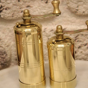 May include: Two gold metal pepper mills with a crank handle. The mills are different sizes and have a shiny finish.