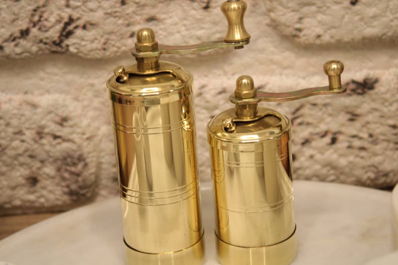 May include: Two gold metal pepper mills with a crank handle. The mills are different sizes and have a shiny finish.