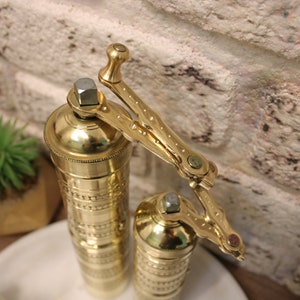 May include: Two gold metal pepper grinders with intricate designs. The grinders have a crank handle with a brass finish.