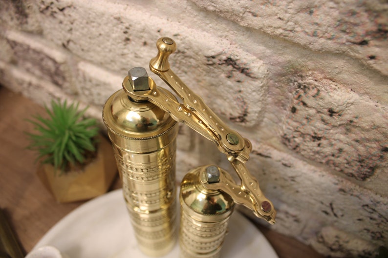 May include: Two gold metal pepper grinders with intricate designs. The grinders have a crank handle with a brass finish.