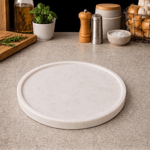 May include: A circular white marble serving tray with a smooth, polished surface and a raised edge. The tray features subtle grey veining throughout the stone, providing a neutral and clean appearance for food presentation or decorative display.