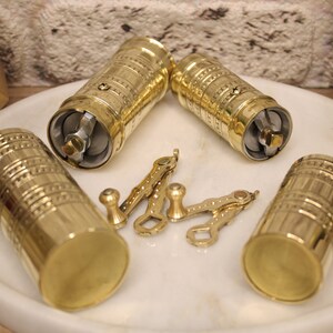 May include: Four gold-colored metal coffee grinders with intricate designs. Each grinder has a handle and a crank. The grinders are on a white surface.