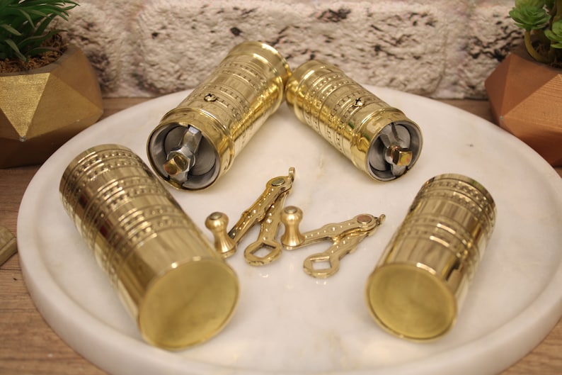 May include: Four gold-colored metal coffee grinders with intricate designs. Each grinder has a handle and a crank. The grinders are on a white surface.
