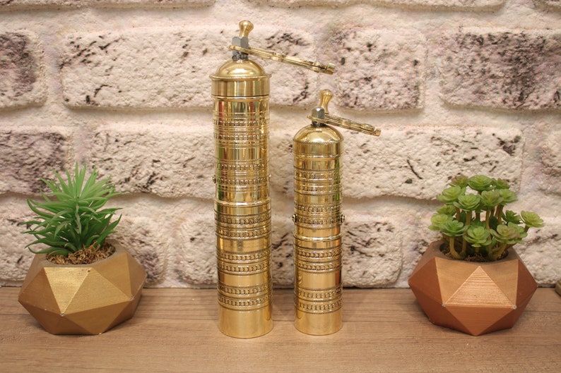 May include: Two gold metal pepper grinders with intricate designs. The grinders are on a wooden surface with two small gold geometric planters with green plants.