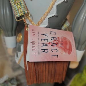 Könnte beinhalten: Eine braune Leder-Umhängetasche mit Fransen. Die Tasche ist mit einem Buch mit dem Titel "The Grace Year" von Kim Liggett verziert. Der Buchumschlag ist rosa mit weißem Text.