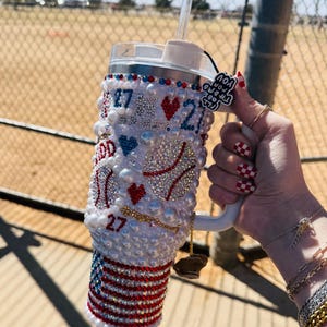 May include: White tumbler with a baseball theme. It is decorated with rhinestones, pearls, and red, white, and blue accents. The tumbler has a straw and a charm that says "You got all the guts to try".